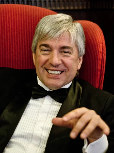 A grey-hair elderly imposing man in an interior. A grey-hair elderly imposing man wearing a jacket sitting in a red armchair and smiling in an old interior.