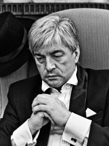 A grey-hair elderly mobster in an interior. A grey-hair elderly mobster in a black suit sitting in a mournful pose against a black-and-white background.