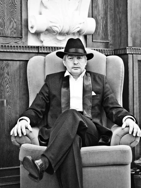 A grey-haired elderly mobster in an interior. A grey-hair elderly mobster wearing a black suit and a hat is sitting in an armchair in an old interior against a black-and-white background.