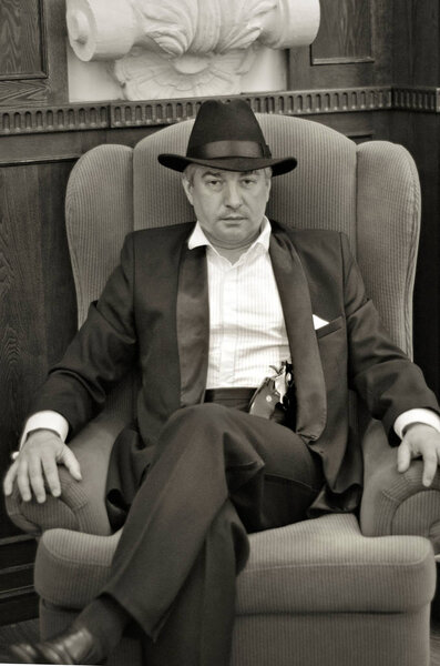 A grey-haired elderly mobster in an interior... A grey-hair elderly mobster with a gun wearing a black suit and a hat is sitting in an armchair in an old interior in retro-style.