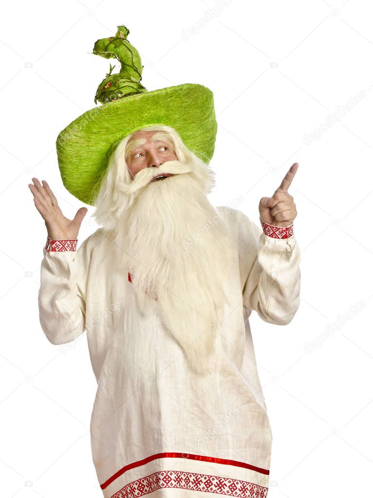 Funny magician with a long gray-haired beard in a green cap points his finger on a white background