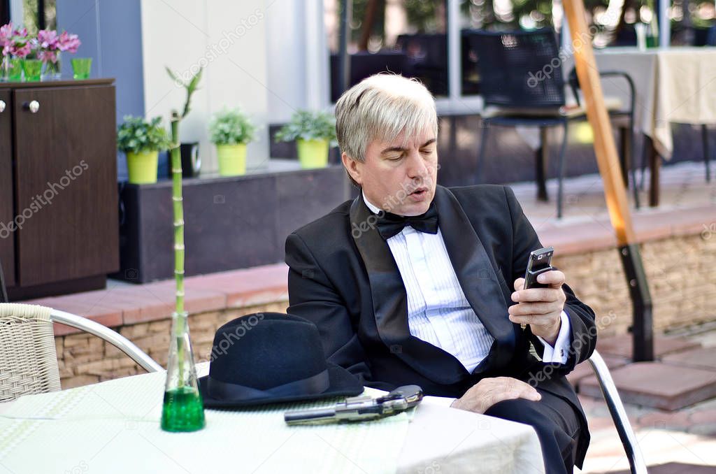 A grey-haired elderly mobster sitting in a summer cafe. Agrey-haired elderly mobster in a black suit with a gun and a mobile phone sitting in a summer cafe.