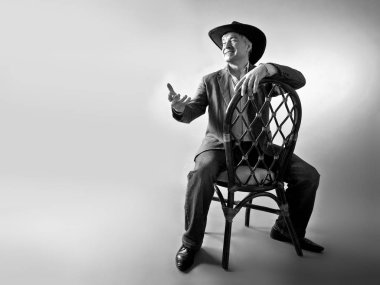 Saygın bir gri saçlı Latin erkek şapka içinde. Saygın bir gri saçlı Latin erkek şapka ve bir ceket bir kovboy gibi bir antika hasır sandalye siyah-beyaz bir arka plan üzerinde oturuyor.