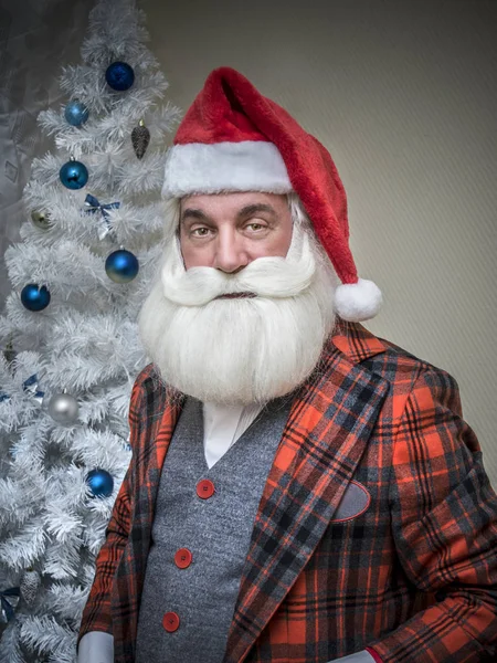 A grey-haired bearded man in an English checkered suit in the image of Santa Claus in a red cap against the background of the Christmas tree.