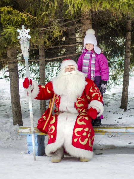 Santa Claus and Father Frost with a girl are sitting on a bench in a public garden.