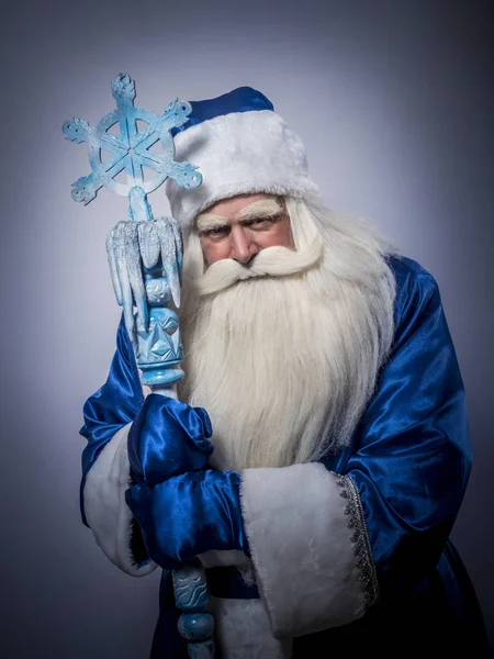 Santa Claus and Father Frost in a blue coat, a hat and with a staff is standing against a dark background.