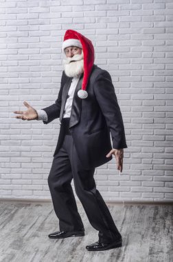Gri saçlı sakallı adam siyah bir takım elbise ve Noel baba resimde kırmızı bere beyaz bir duvar duran ve duygusal elleriyle gesticulating.