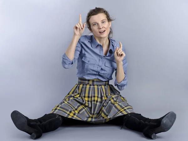 A beautiful European woman is funny and originally sits on the floor. A beautiful European woman is funny and originally sits on the floor in a blue shirt and a checkered skirt on a blue background.