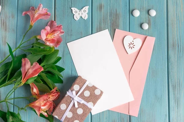 Mock up blank paper, mail envelope on a blue wooden background with natural flowers of pink color and butterflies. Blank, frame for text. Greeting card design with flowers. Aalstroemeria on wooden bac