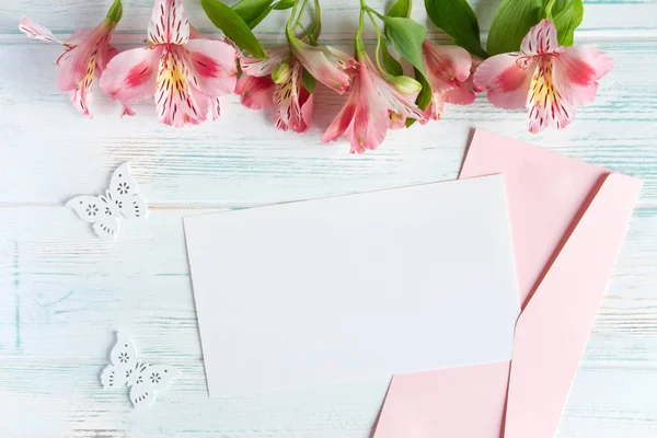Finge papel en blanco y sobre sobre fondo blanco de madera con flores naturales de color rosa. En blanco, marco para texto. Diseño de tarjeta de felicitación con flores. Aalstroemeria. Piso tendido, vista superior . — Foto de Stock