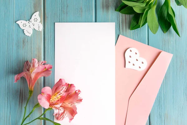 Simular papel en blanco, sobre de correo sobre un fondo de madera azul con flores naturales de color rosa y mariposas. En blanco, marco para texto. Tarjeta de felicitación con flores. Aalstroemeria sobre fondo de madera — Foto de Stock