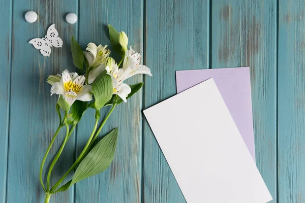 Mock up blank paper, mail envelope on a blue wooden background with natural white flowers and butterflies. Blank, frame for text. Greeting card with flowers. Aalstroemeria on wooden background.