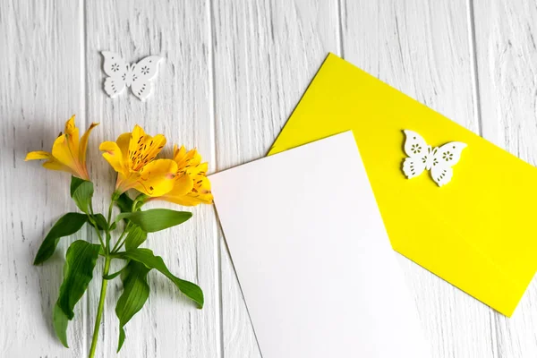 Mock up blank paper, mail envelope on a white wooden background with natural flowers of yellow color and butterflies. Frame for text. Greeting card design with flowers. Aalstroemeria background. — Stock Photo, Image