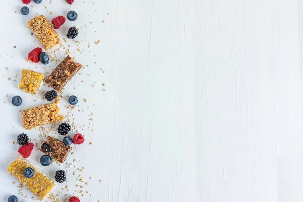 Frame for text with granola and natural berries of raspberry, blackberry, blueberry and oatmeal on a white wooden background. Useful cereal bars for snacking and breakfast
