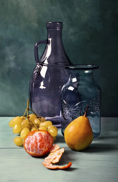 Still life with blue glass vase, lilac glass jug and fruit on turquoise background. Tinted