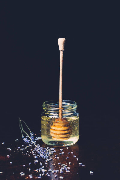 selective focus of jar with honey stick and petals of lavender on black background 