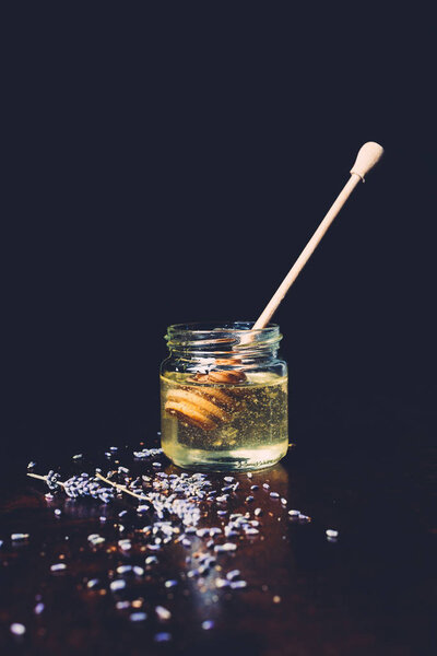 close up view of jar with honey stick and petals of lavender on black background 