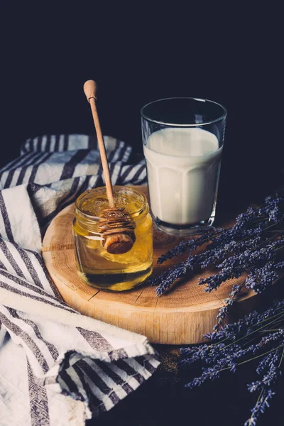 Selective focus of glass of milk, jar with honey stick and lavender on black background — Stock Photo