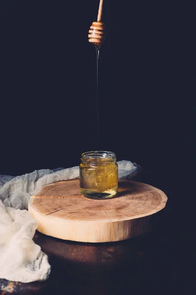 Selective focus of honey stick with flowing honey into jar on wooden slice on black background — Stock Photo