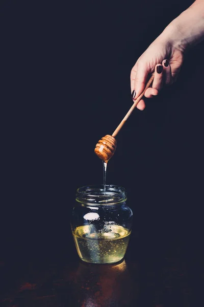 Partial view of woman putting honey stick into glass with honey on black background — Stock Photo