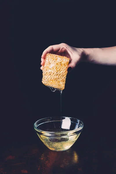 Cropped image of woman holding honeycomb with flowing honey into bowl on black background — Stock Photo