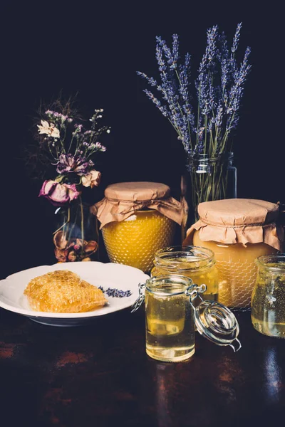 Selective focus of jars with honey, dried flowers, lavender and honeycomb on black background — Stock Photo