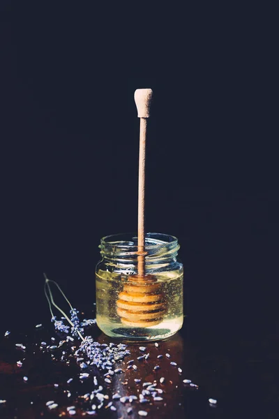 Selective focus of jar with honey stick and petals of lavender on black background — Stock Photo
