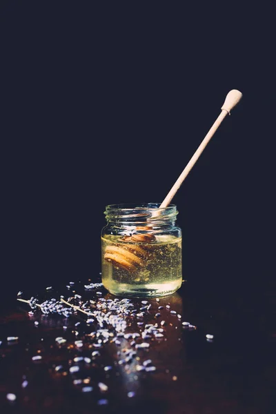 Close up view of jar with honey stick and petals of lavender on black background — Stock Photo