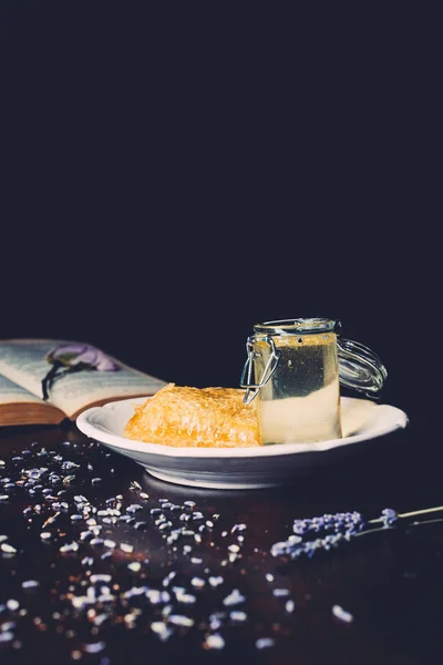 Selective focus of plate with jar and honeycomb surrounded by lavender petals on black background — Stock Photo