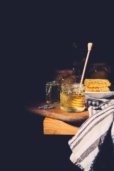 Selective focus of honey stick in jar with honey on cutting board at table on black background — Stock Photo