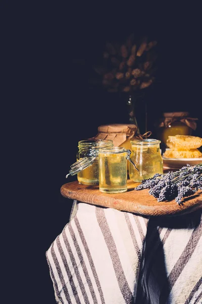 Selective focus of cutting board with different jars of honey and lavender on black background — Stock Photo