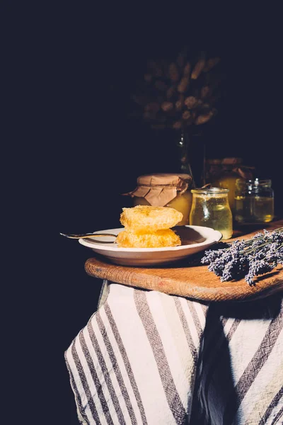 Selective focus of plate with honeycombs, jars with honey and lavender on black background — Stock Photo