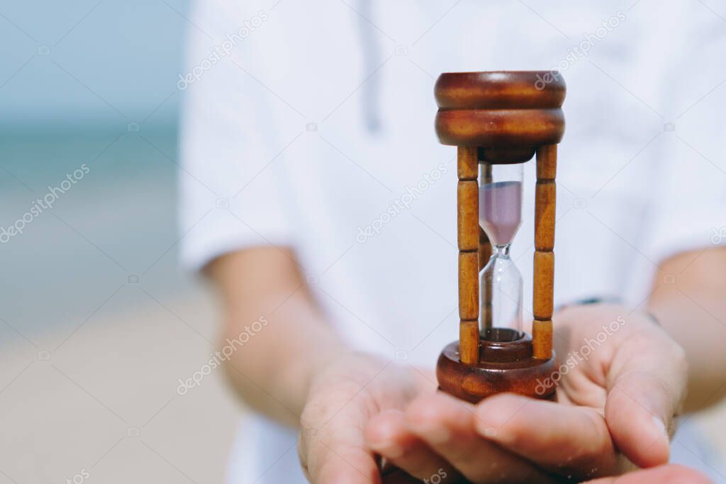 Hand hold small hourglass model show time is flowing on summer beach background.