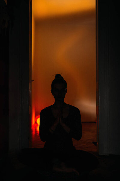 Silhouette of young woman yoga instructor meditating sitting on a rug on the floor in a black solid suit getting ready for a great workout. Toned image.