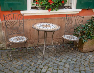 İki eski mozaik sandalye ve evin önünde yuvarlak bir mozaik masa.