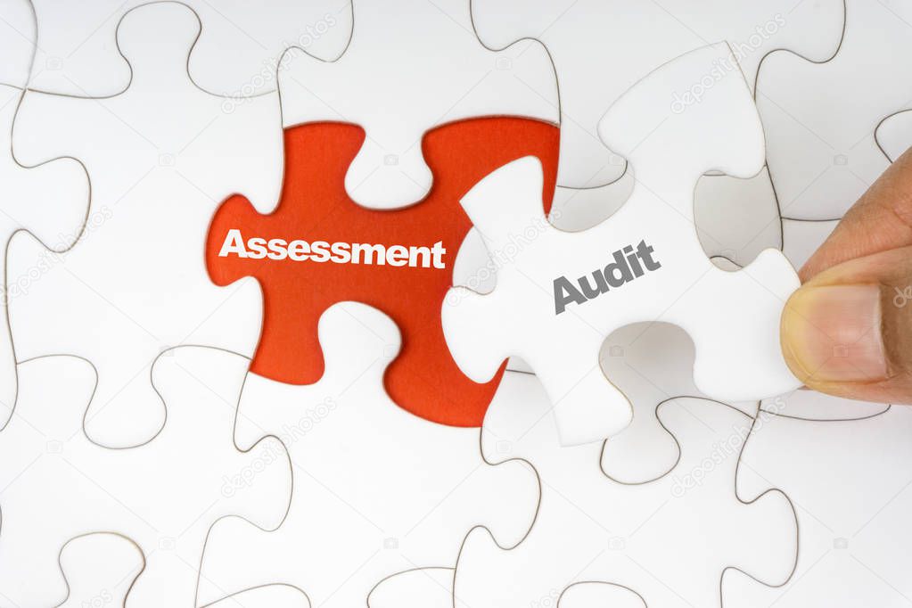Hand holding piece of jigsaw puzzle with word AUDIT ASSESSMENT. Selective focus
