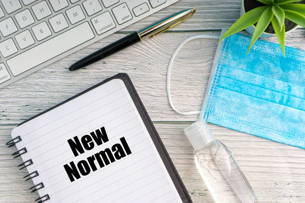 NEW NORMAL text with notebook, safety face mask, keyboard, fountain pen, hand sanitizer and decorative plant on wooden background. Covid 19 and coronavirus concept