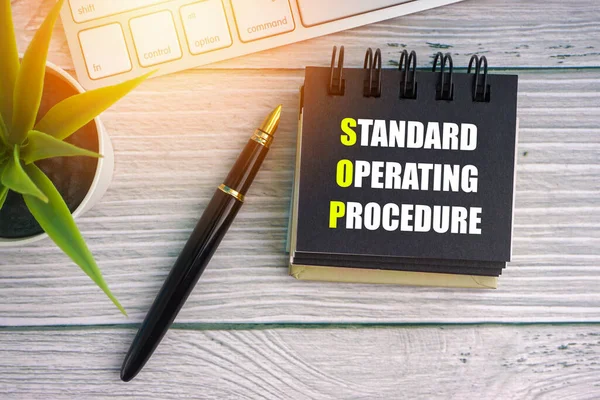 Dolma kalem, dekoratif bitki, klavye ve ahşap arka planda not defteri ile STANDARD OPERATING PROCEDURE metni. İş konsepti