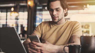Kafede cep telefonu ve dizüstü bilgisayar kullanan genç adam.