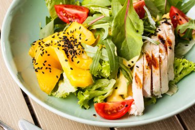 Ahşap arka plan üzerinde beyaz bir tabak üzerinde ızgara tavuk, mango, marul, avokado, domates, arugula, peynir sause ile Salata