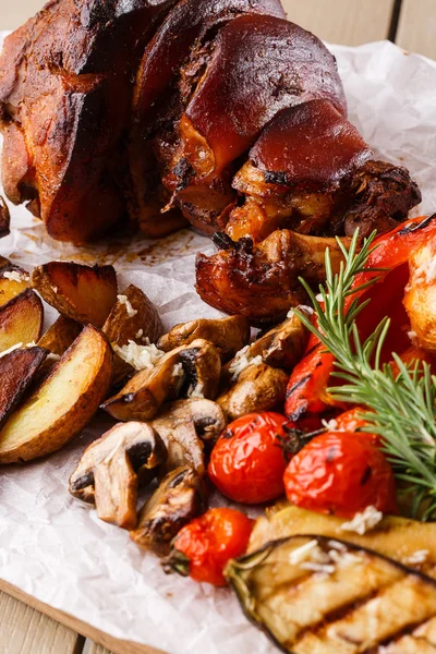Grilled pork knuckle with grilled tomatoes, champignons, vagetable marrow, eggplant, red sweet pepper and baked potatoes and parchment on a wooden cutting board. top view — Stock Photo, Image