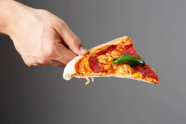 Slice of Delicious hot homemade pizza in hand on gray background. Pizza - Fresh homemade pizza with pepperoni, cheese and tomato sauce, tomatoes, sweet corn and basil with copy space. — Stock Photo, Image