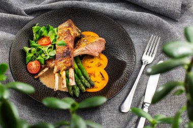 Kuşkonmaz, sarı domates ve kuzu sosuyla servis edilen kızarmış kuzu pirzolası. Siyah taş tabakta çatal ve bıçakla servis edilen fileto kuzu eti. Gri arka plan. Ton balığı salatası. Yukarıdan yatay görünüm.