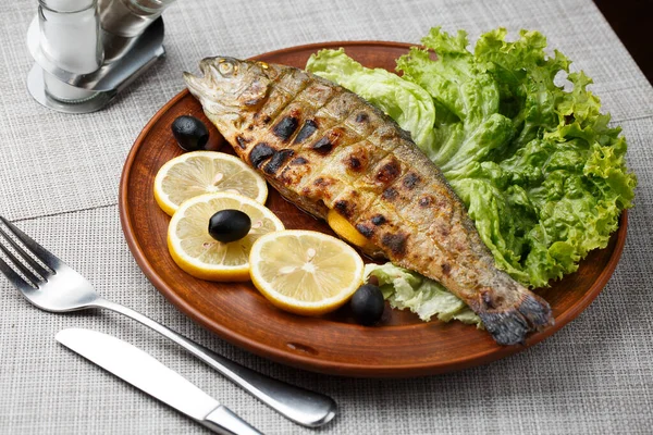 Grilled trout in brown clay bowl close-up. Front view of plate with grilled trout garnishe with salade, lemon and olives. Focus on fried fish. Grey background. horizontal photo — Stock Photo, Image