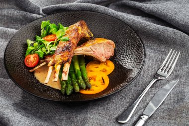 Kuşkonmaz, sarı domates ve kuzu sosuyla servis edilen kızarmış kuzu pirzolası. Siyah taş tabakta çatal ve bıçakla servis edilen fileto kuzu eti. Gri arka plan. Ton balığı salatası. Yukarıdan yatay görünüm.