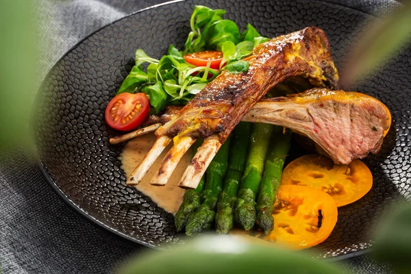 Roasted rack of lamb served with asparagus, yellow tomatoes and lamb sauce. Loin lamb Served in black stone plate with fork and knife. Gray background. Tuna salad. Top view from above horizontal. — Stock Photo, Image