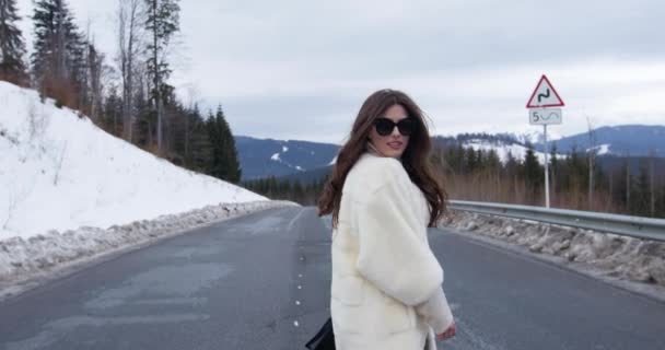 Powrót Zobacz dziewczynę spacerującą po drodze na zimowym tle górskim. Czarująca młoda kobieta w stylowym swetrze, białym modnym futrze i okularach przeciwsłonecznych. Futro i koncepcja mody. Piękni ludzie. 4k — Wideo stockowe