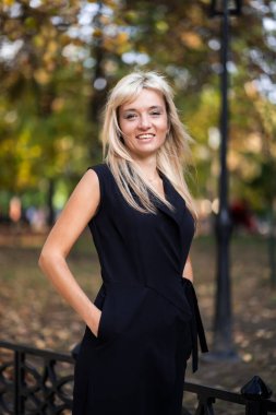 Sonbahar modası açık havada. Havalı siyah tulumlu sarışın kız, parktaki bulanık sarı-yeşil ağaçların arka planında sonbahar yaşam tarzı. Dikey