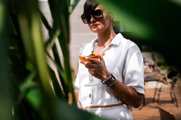 Kısa esmer saçlı, beyaz elbiseli ve güneş gözlüklü güzel bir kadın. Moda sokak fotoğrafçılığı. Ahşap arka planda aperol kokteyli ile poz veren manken.. — Stok fotoğraf