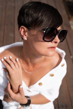 Kısa esmer saçlı, beyaz elbiseli ve güneş gözlüklü güzel bir kadın. Moda sokak fotoğrafçılığı. Moda modeli kahverengi ahşap arka planda oturuyor ve rahatlıyor. Güneş gözlüğü dükkanı